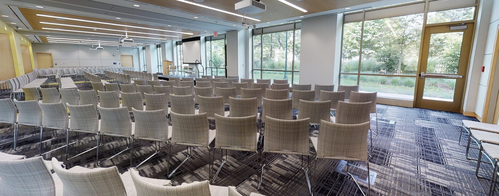 A meeting room at the East Campus Commons, set with rows of upholstered chairs facing a projection screen, with floor-to-ceiling windows along one wall looking out onto trees and lawn.